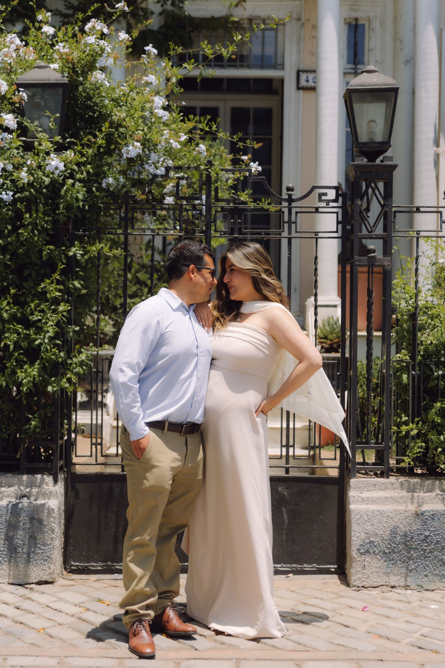 Couple leaning in for a kiss outside elegant white building with black iron gate and flowering shrubs.