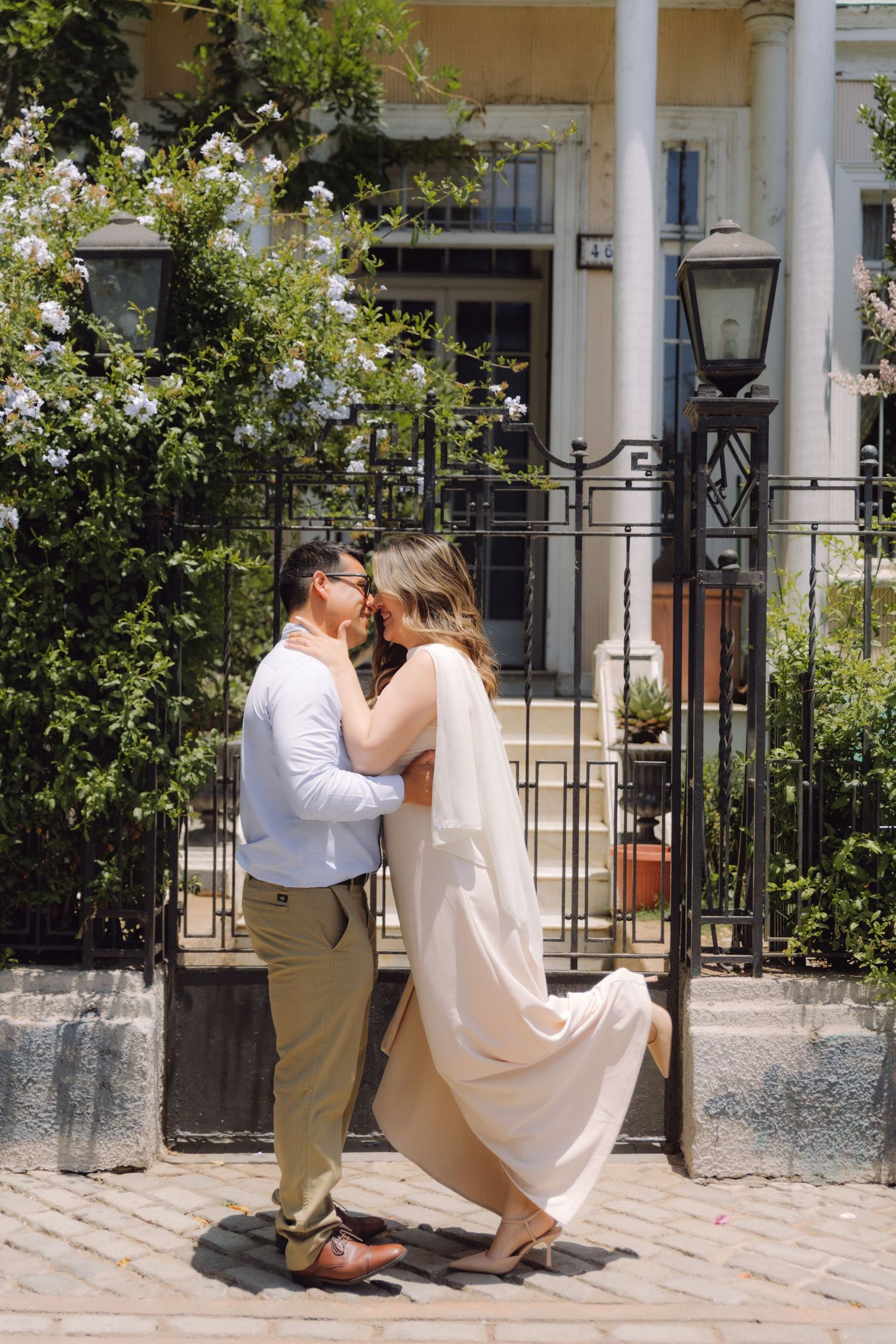 Couple embracing and kissing on cobblestone street in front of white colonial building with blooms.