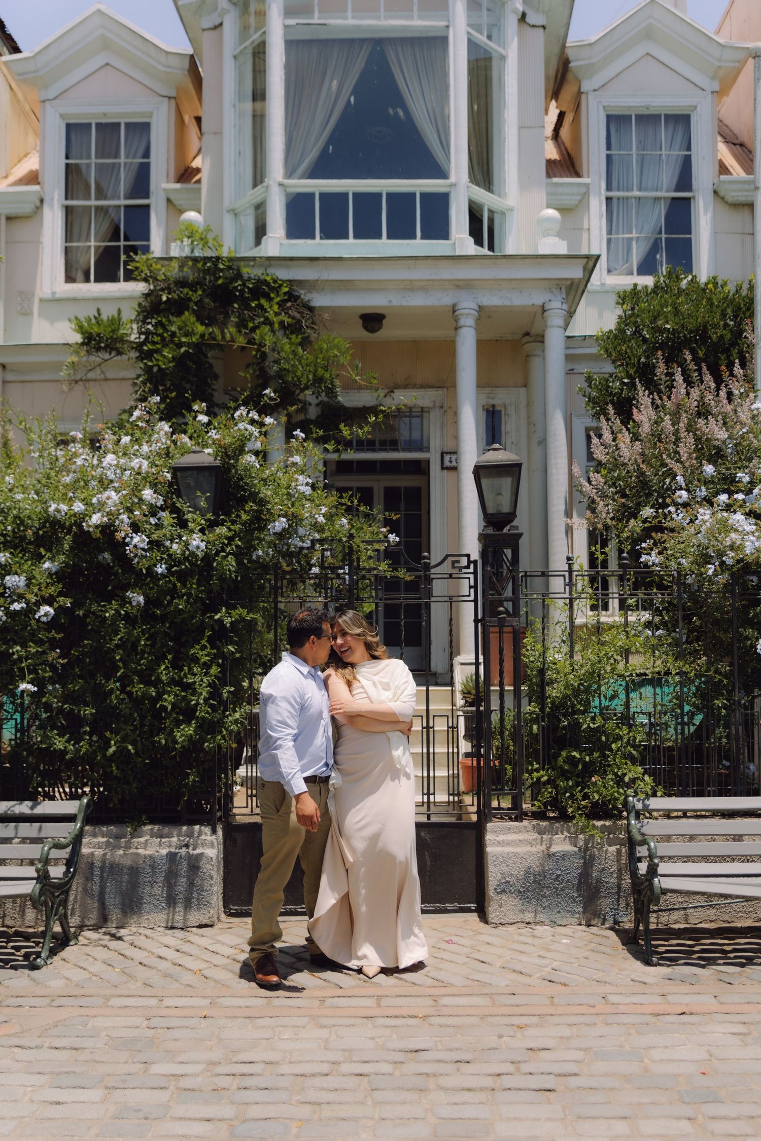 Couple sharing intimate moment on cobblestone path before grand white Victorian home with lush garden.