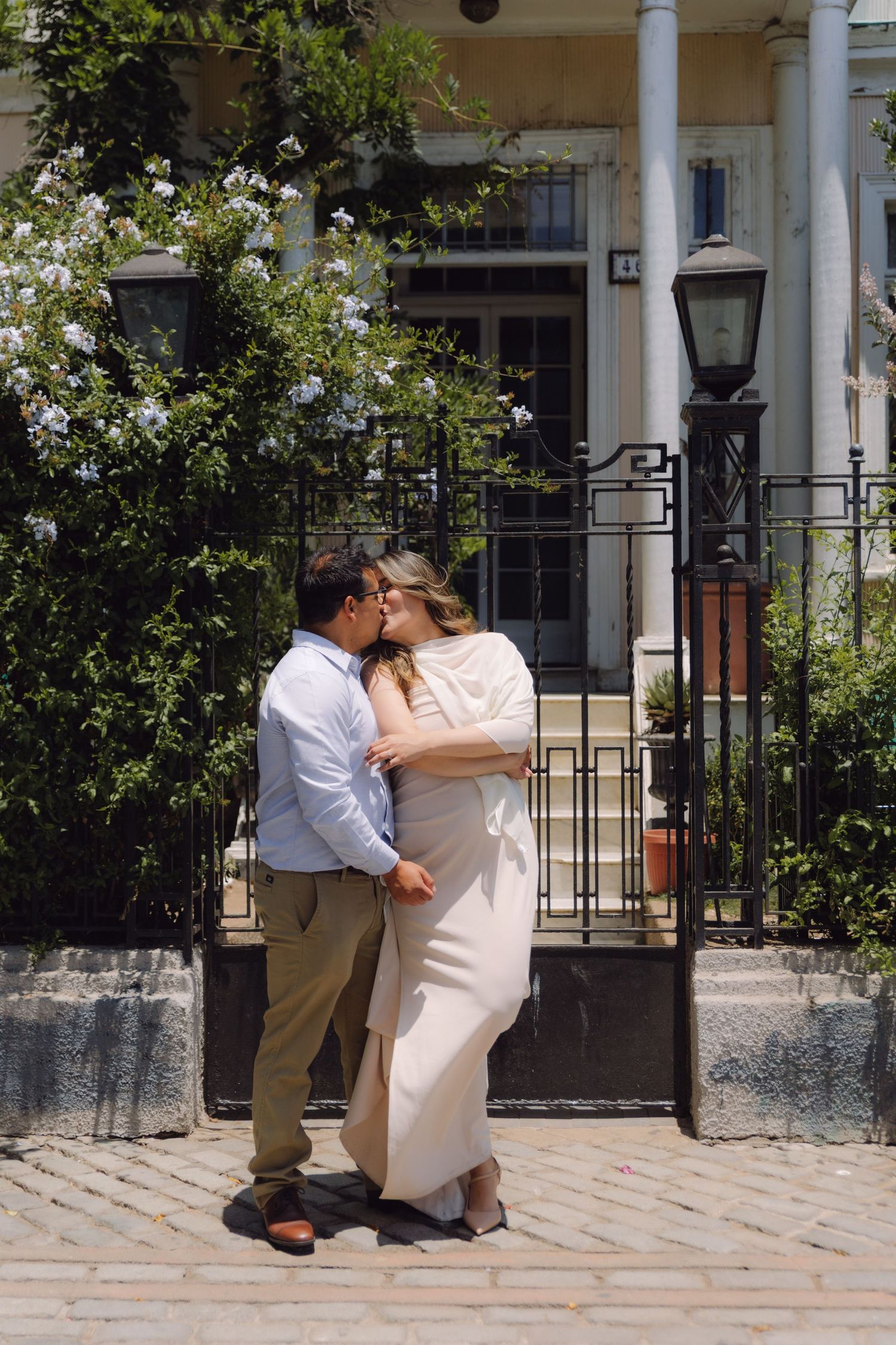 Couple kissing tenderly outside elegant white building with iron gate and blooming white flowers.