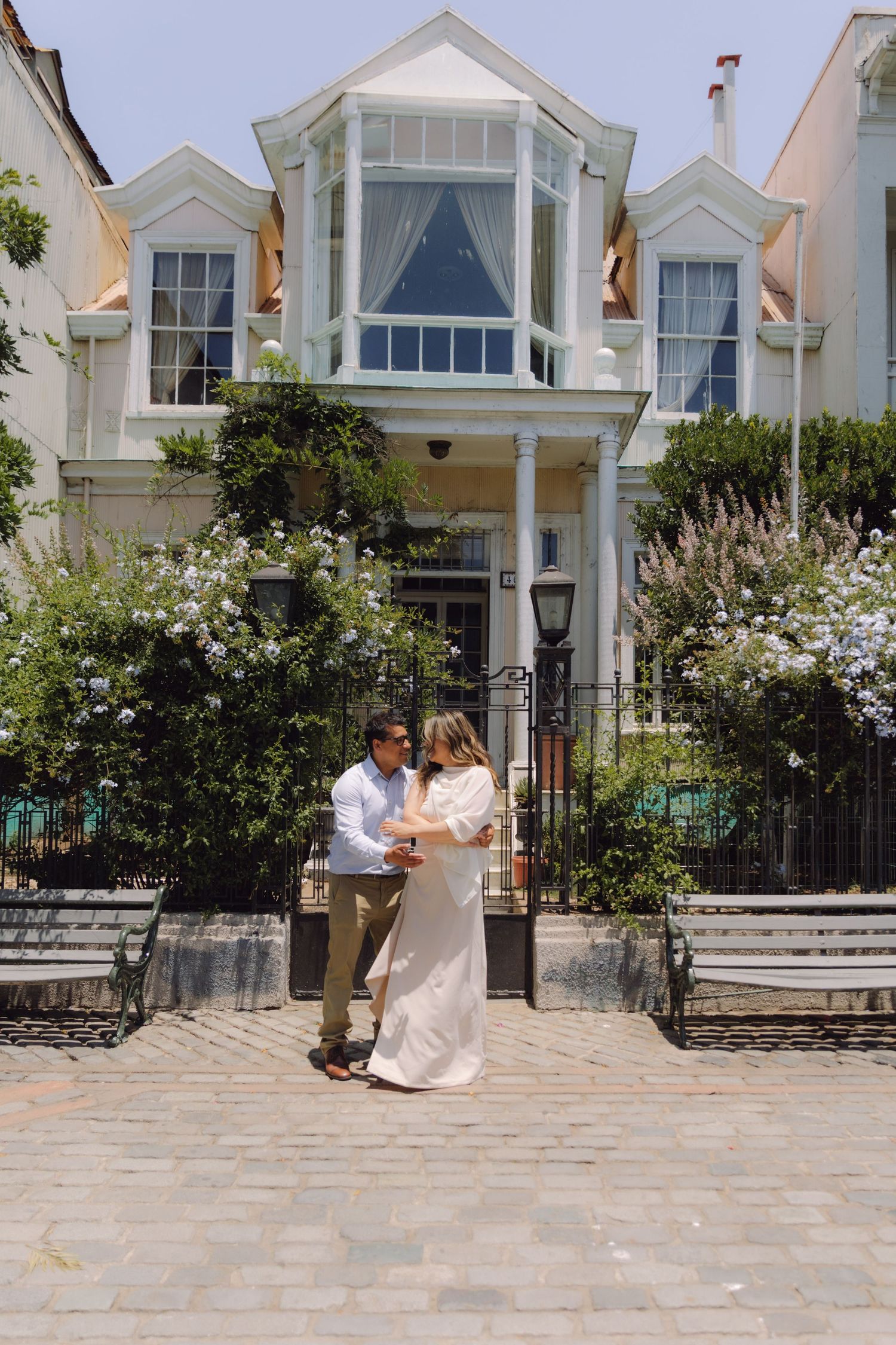 Couple embracing in front of a charming Victorian house with blooming white flowers on a sunny day.