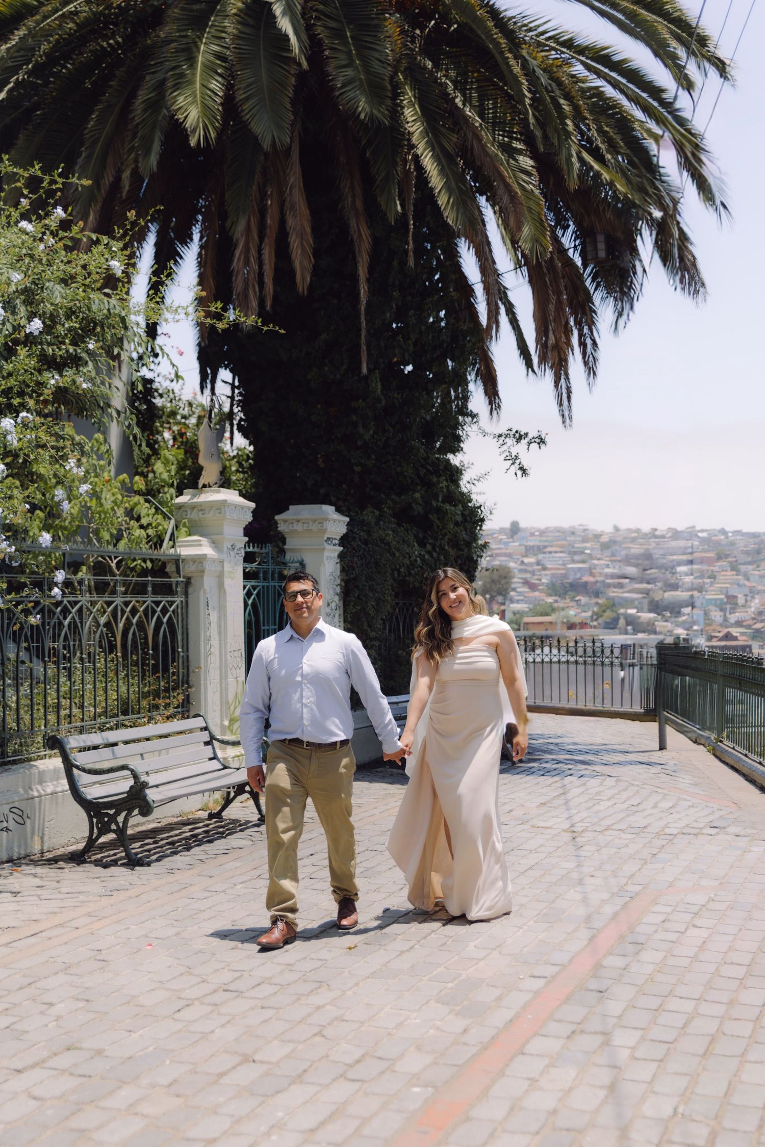 Couple holding hands and walking on a cobblestone terrace with a palm tree and city view behind them.