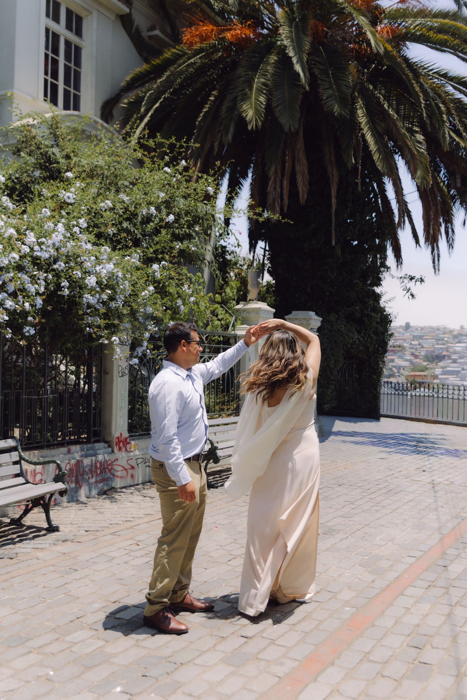 Man twirls woman in elegant white dress near a palm tree and historic building on a sunny day.