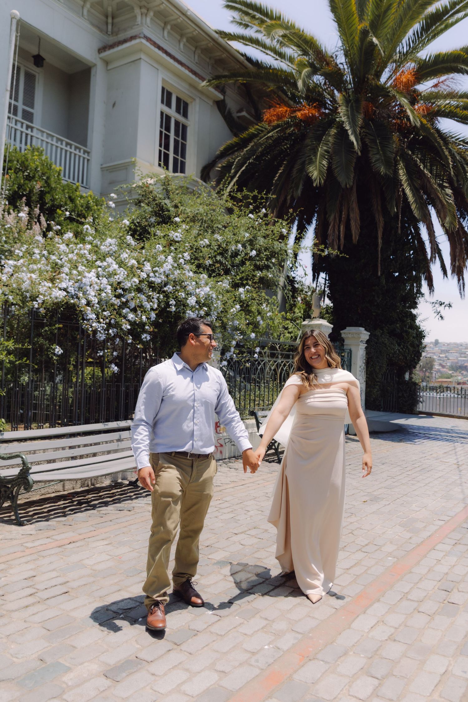 Happy couple walks hand in hand past a palm tree and blooming garden on a sunny cobblestone plaza.