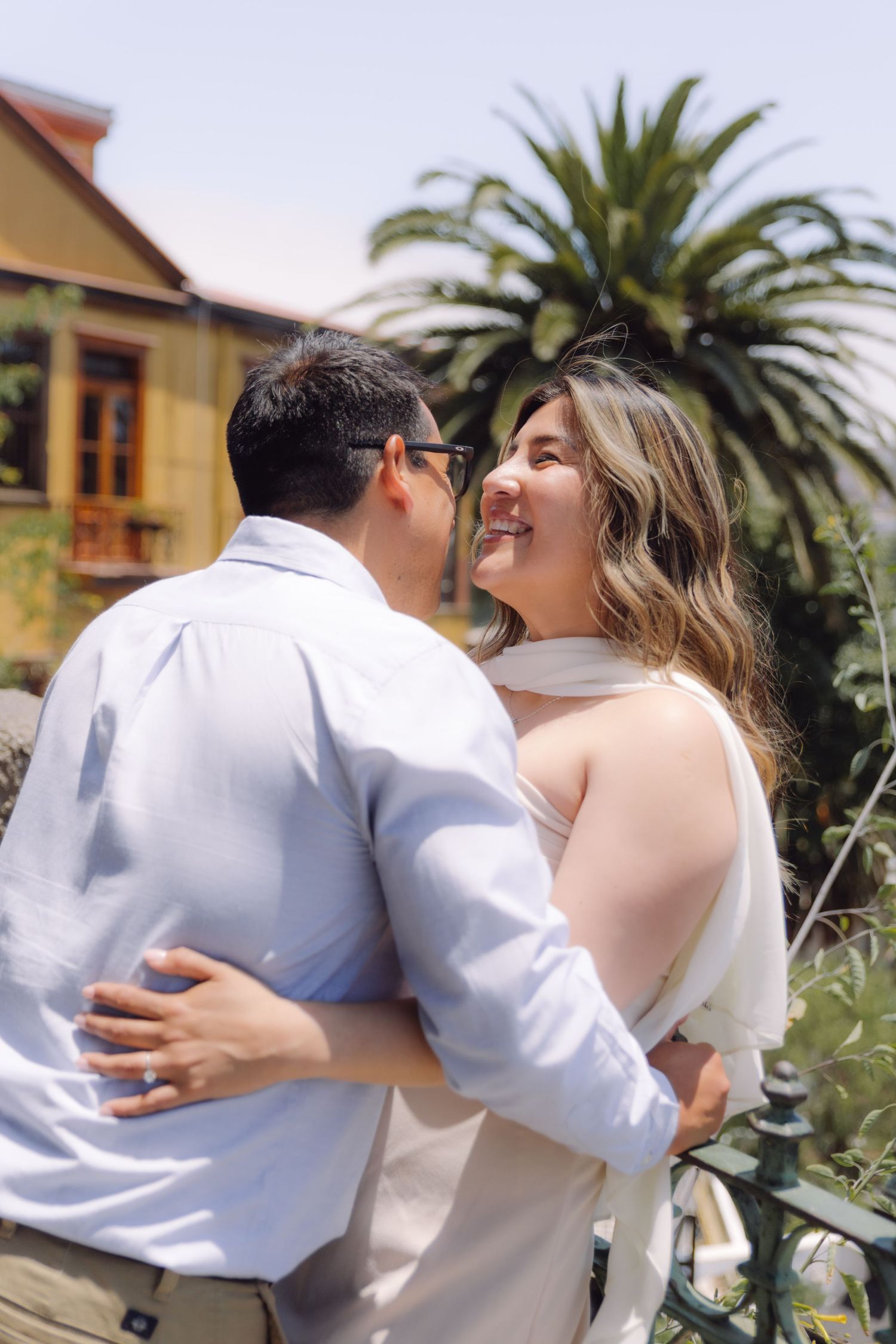 Smiling couple embracing outdoors near tropical palm trees and colorful building on sunny day.
