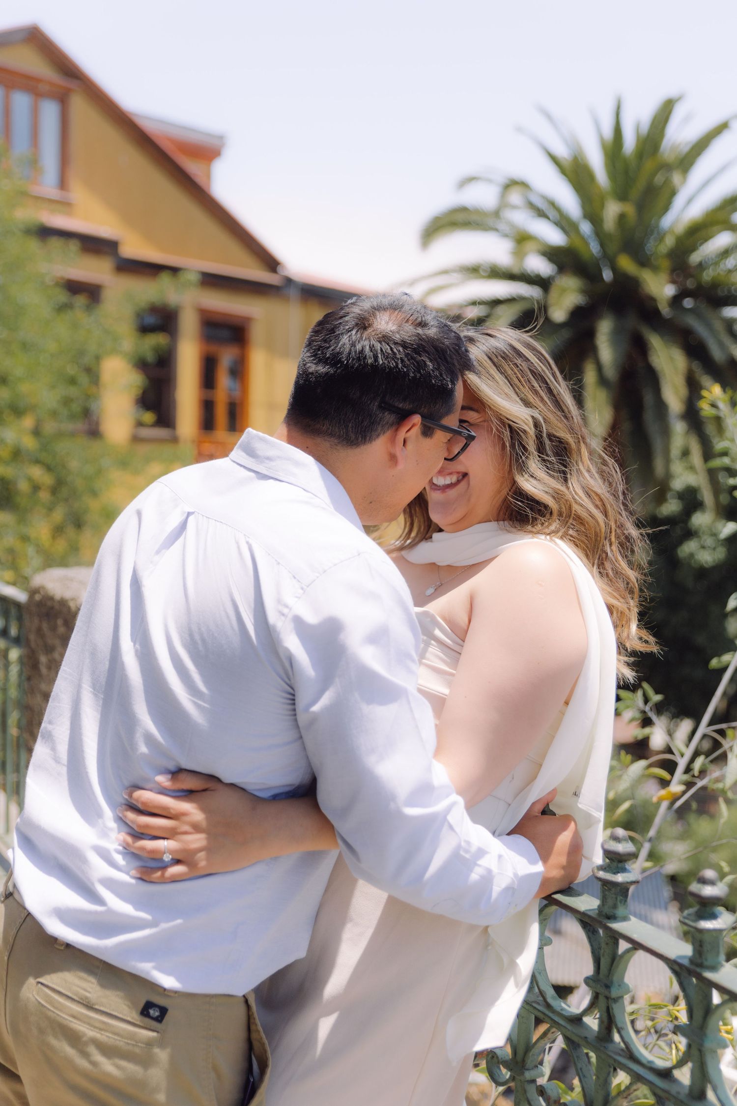 Happy couple sharing romantic moment outdoors with palm trees and yellow building in background.