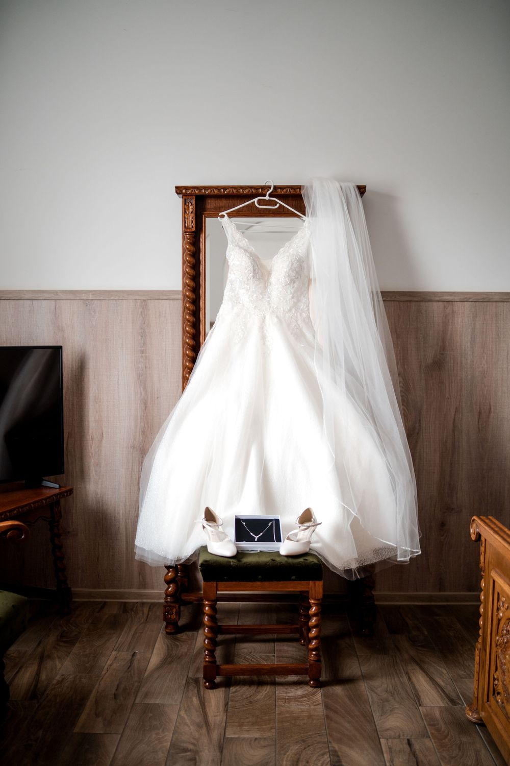 White lace ball gown wedding dress hanging on ornate wooden mirror frame with veil and bridal shoes below.
