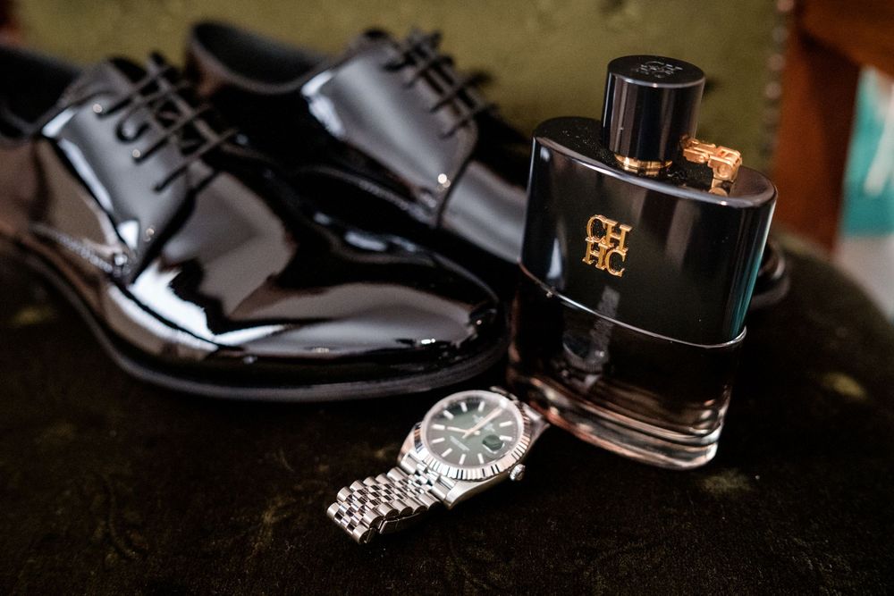 Shiny black patent leather dress shoes, Carolina Herrera cologne bottle, and a silver Rolex watch arranged on a dark surface.