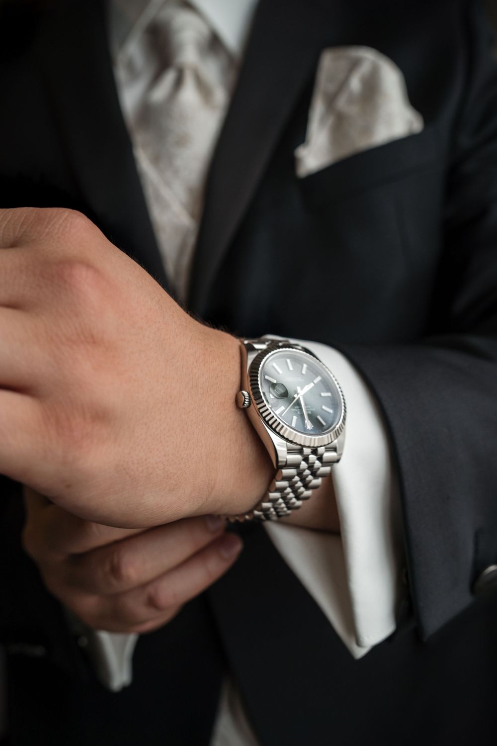 Man in black suit adjusting silver Rolex Datejust watch with blue dial, wearing white dress shirt and silk tie.