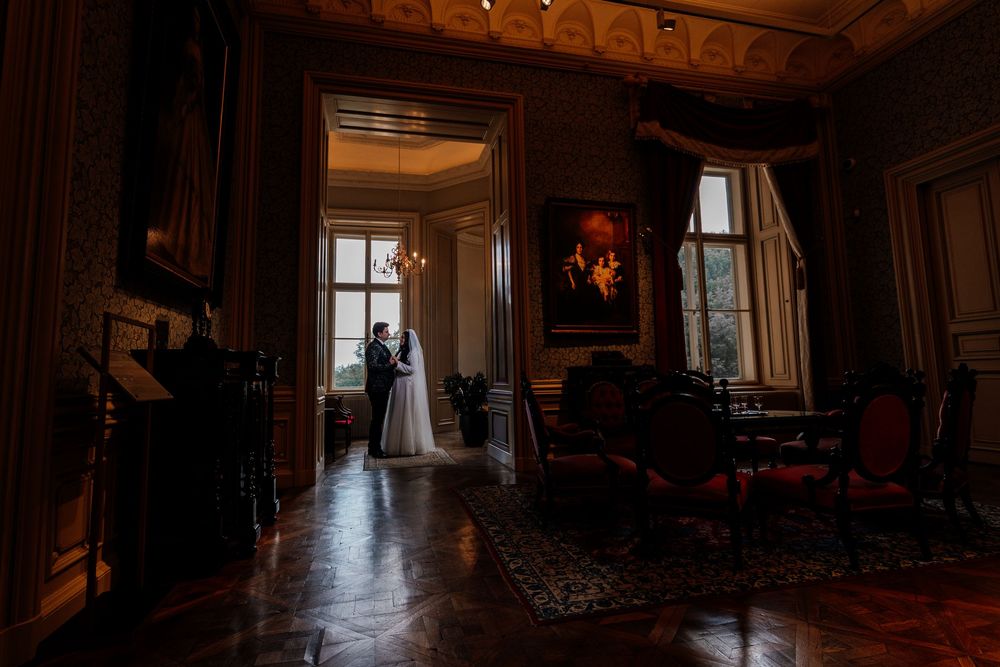 Bride and groom silhouetted in a grand, dimly lit historic mansion interior with ornate decor and parquet floors.
