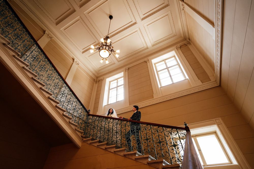 Two people stand on an ornate staircase in an elegant historic building with chandelier and tall windows.