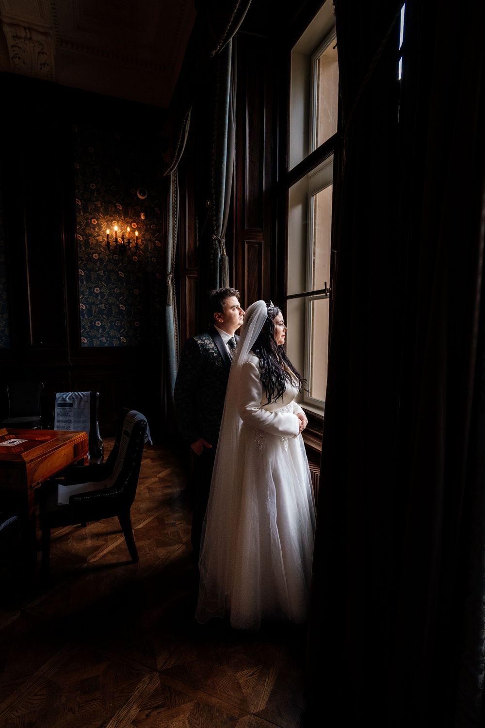 Bride and groom standing by a large window in a dark elegant room, sharing a romantic moment on their wedding day.