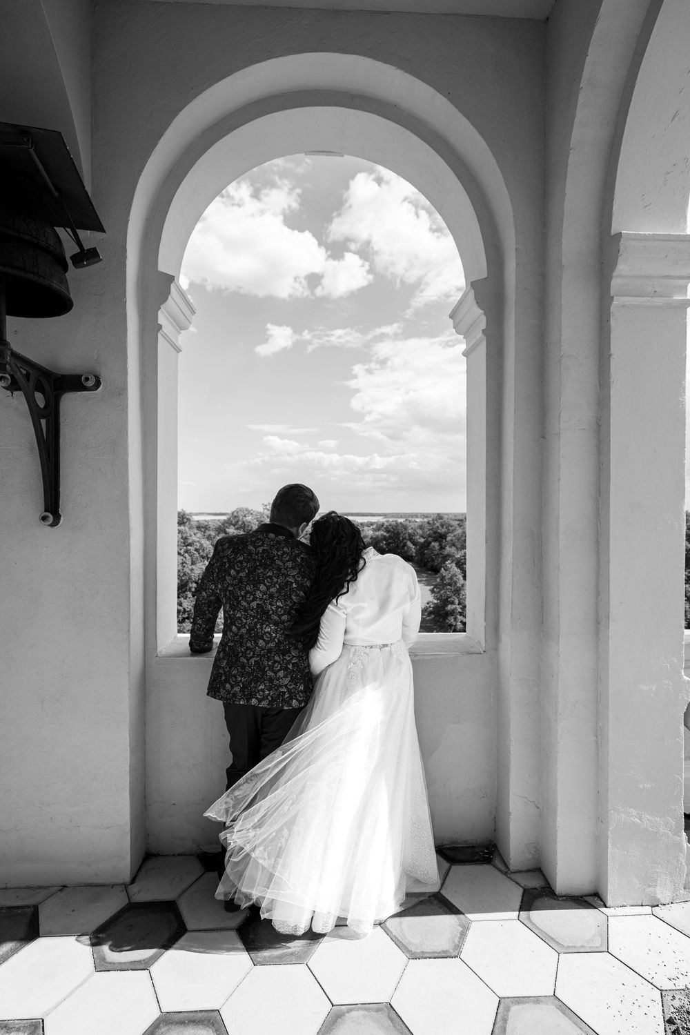 Black and white photo of a bride and groom embracing under an arch, gazing at a scenic landscape on their wedding day.