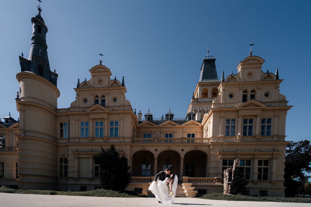 Bride and groom pose in front of a grand baroque palace with ornate facade under a clear blue sky.