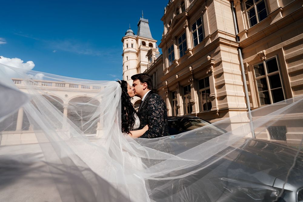 Bride and groom share a romantic kiss outdoors with flowing veil and elegant historic building backdrop on sunny day.