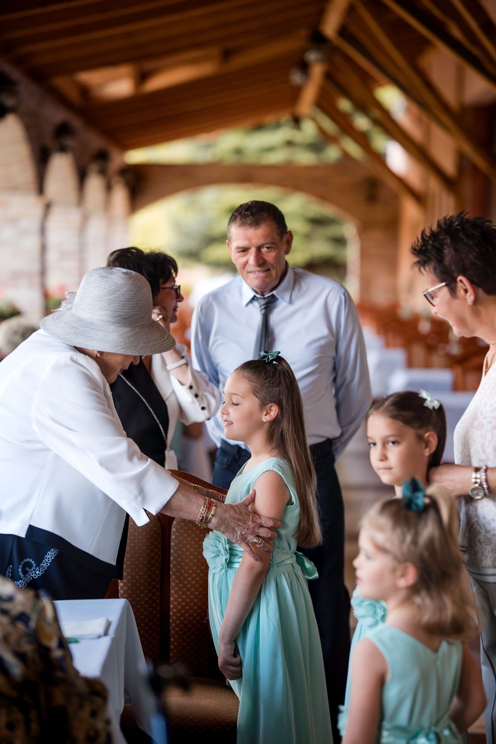 Elderly woman in white hat interacting with girls in teal dresses at a wedding reception under a wooden pavilion.