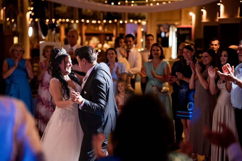 Bride and groom share first dance at wedding reception surrounded by clapping guests under warm string lights.