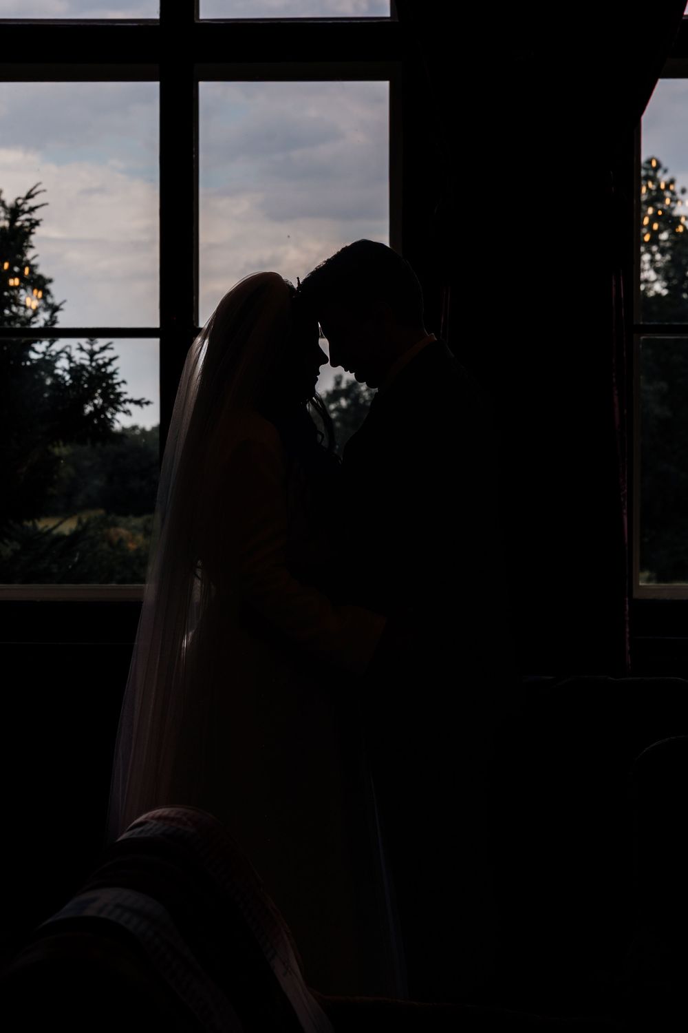 Silhouette of bride and groom touching foreheads by a large window at dusk, with soft evening light and veil flowing.