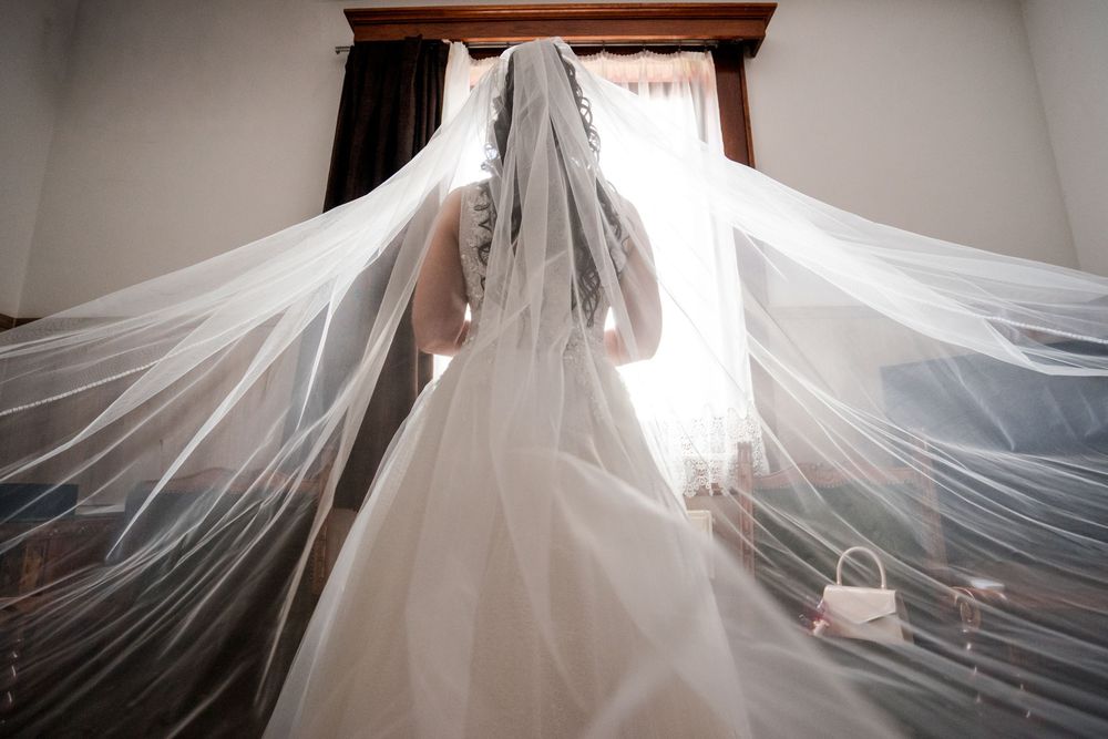 Bride in white wedding gown and flowing veil seen from behind, standing before a bright window.
