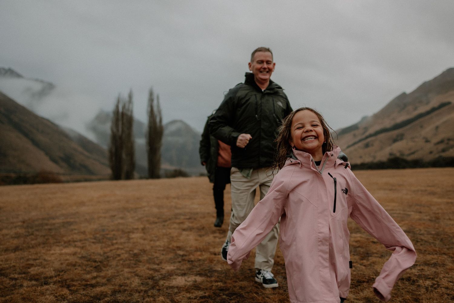 A joyful girl in a pink jacket runs outdoors with a smiling man in a green jacket, set against misty mountains.