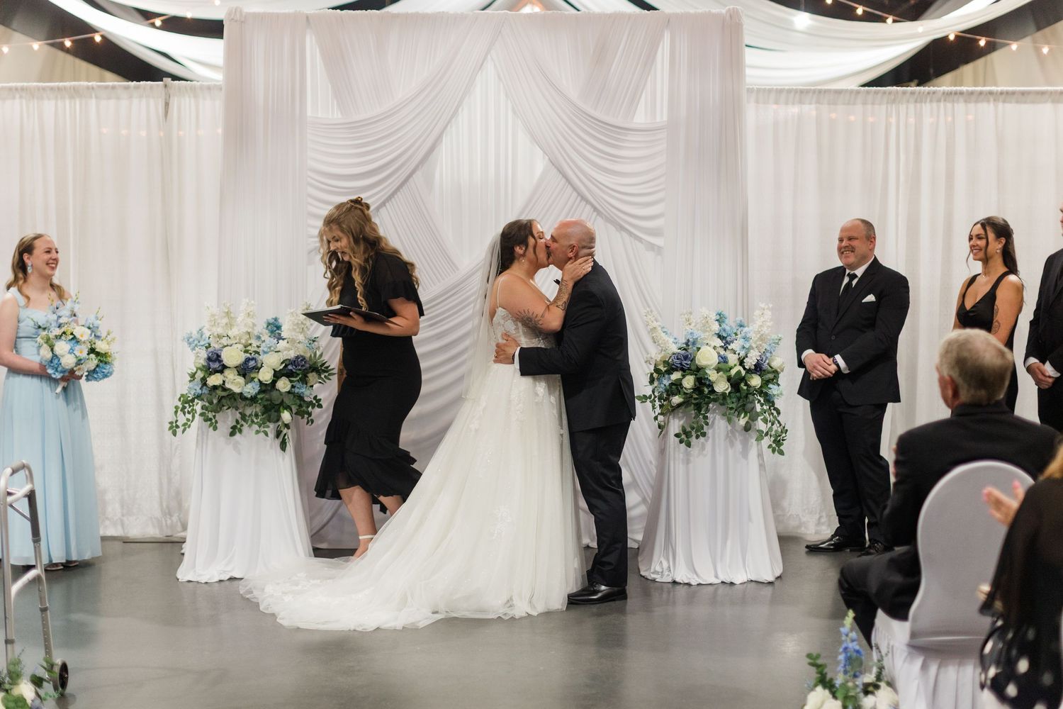 Bride and groom share first kiss at elegant wedding ceremony with white draping and blue floral arrangements.