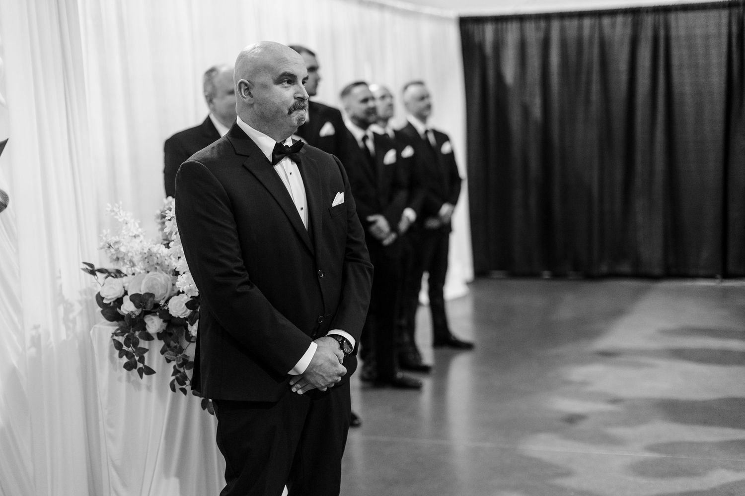Groom in black tuxedo stands at wedding altar with groomsmen behind him in black and white photo.