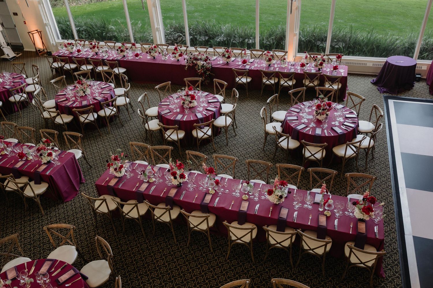 Elegant wedding reception venue with burgundy and pink table settings, round and rectangular tables with cross-back chairs.
