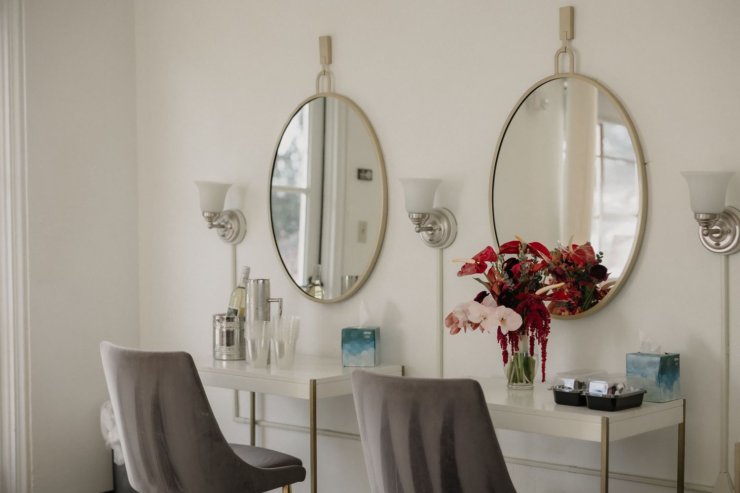 Elegant vanity table with two oval mirrors, floral arrangement, and gray velvet chairs in a bright white room.