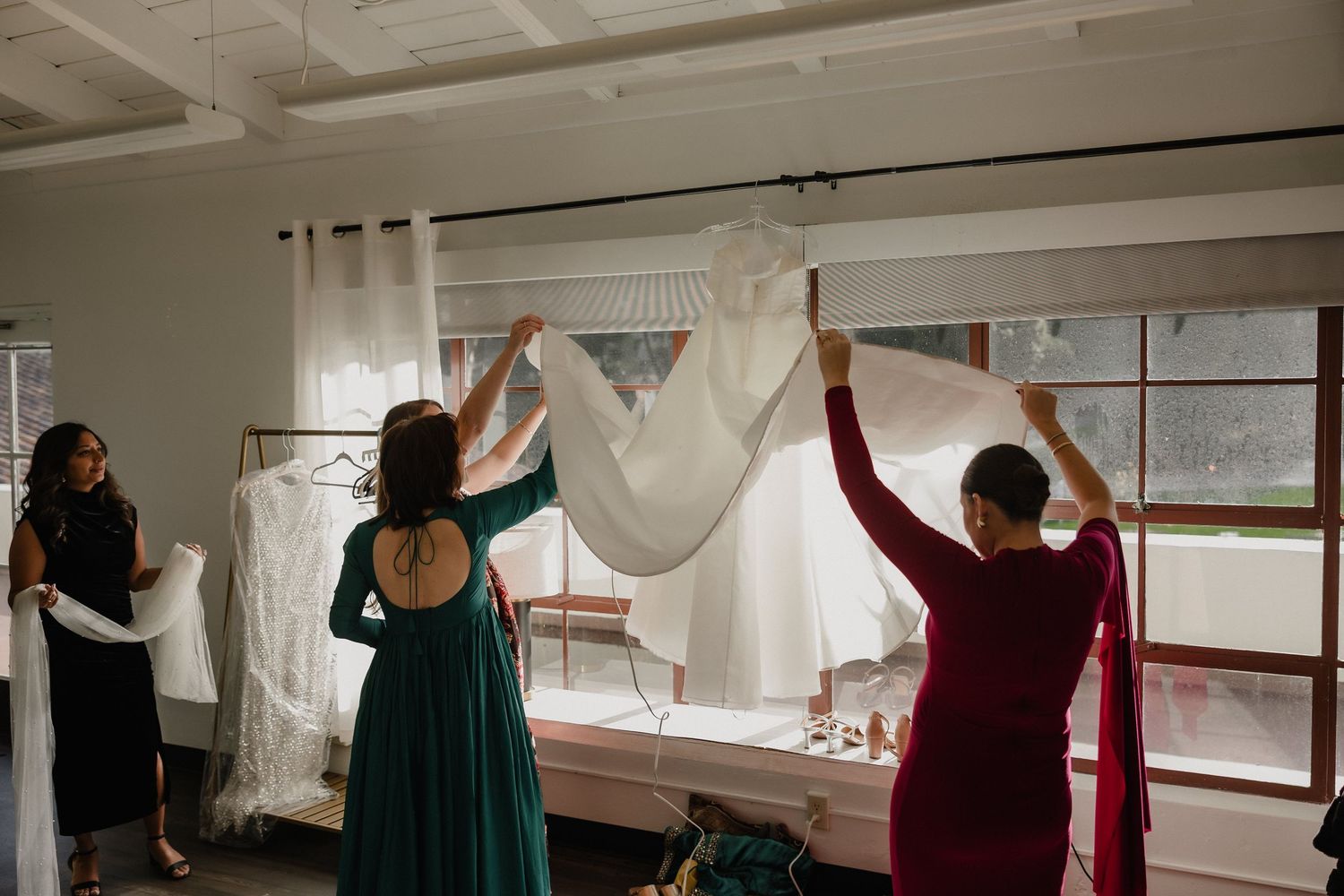 Bridesmaids in green and red dresses holding up a white wedding veil near bright windows.