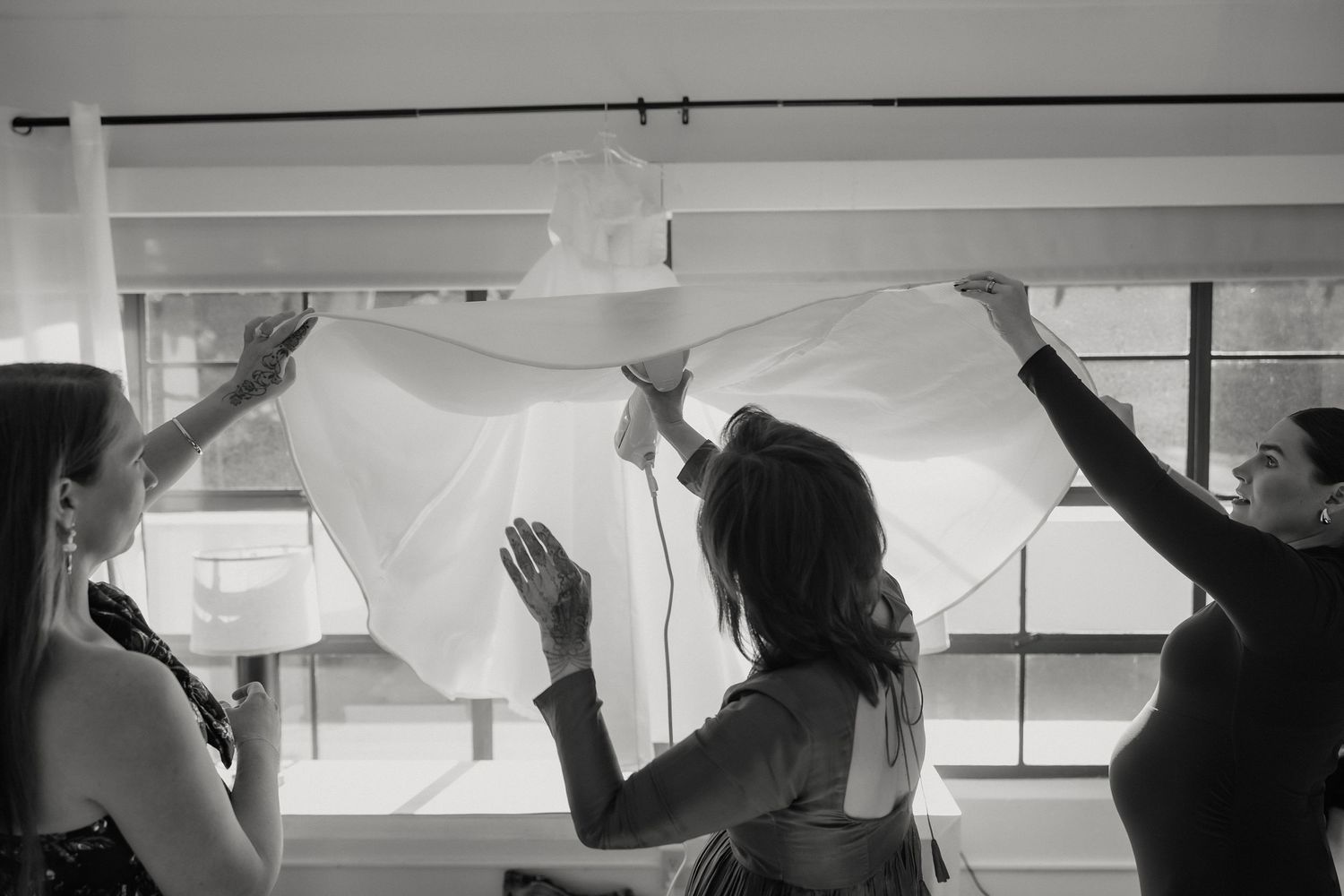Black and white photo of women lifting a flowing white wedding veil near large windows.