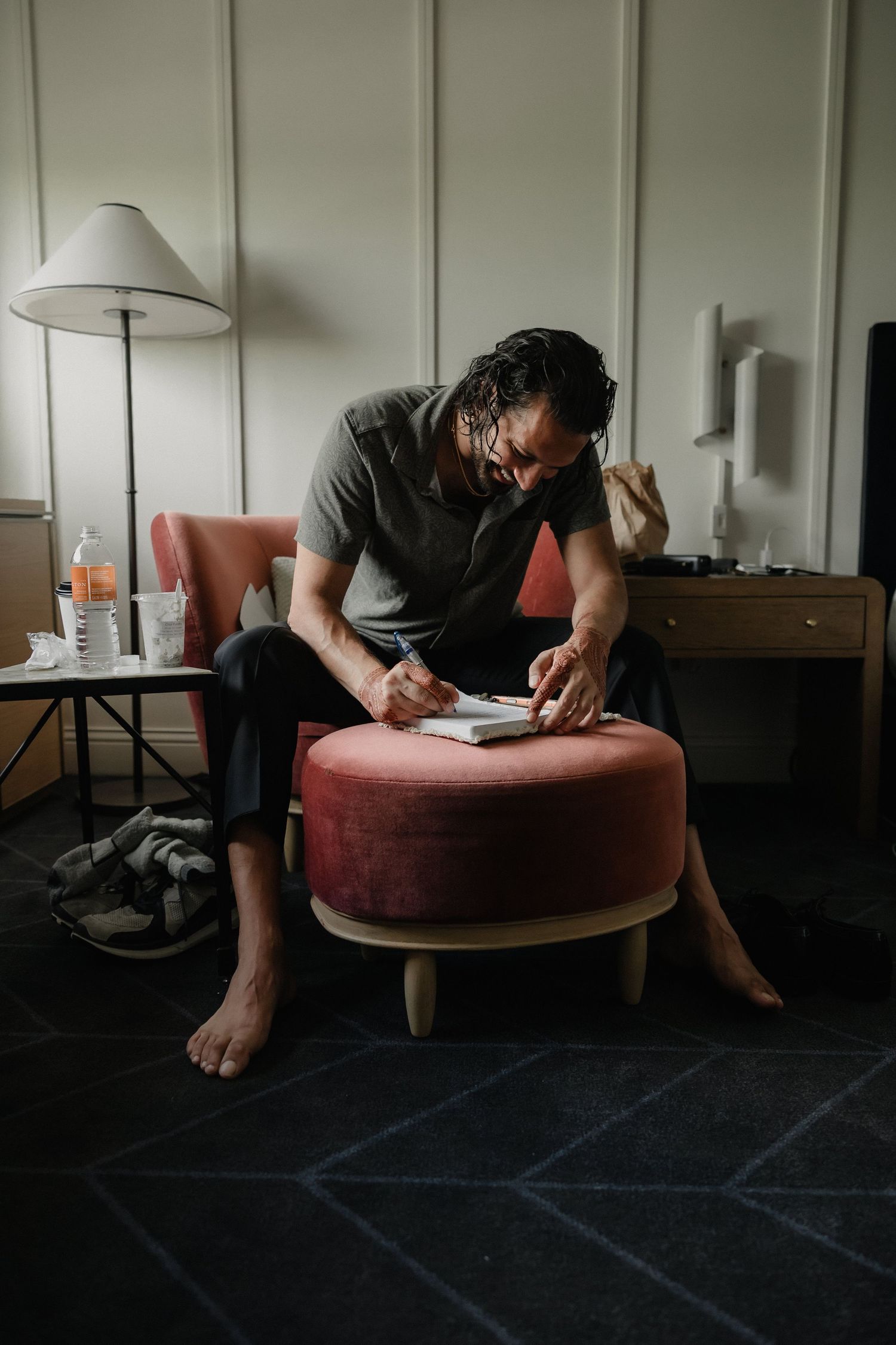 Man sits in a pink chair writing in a notebook in a dimly lit hotel room.