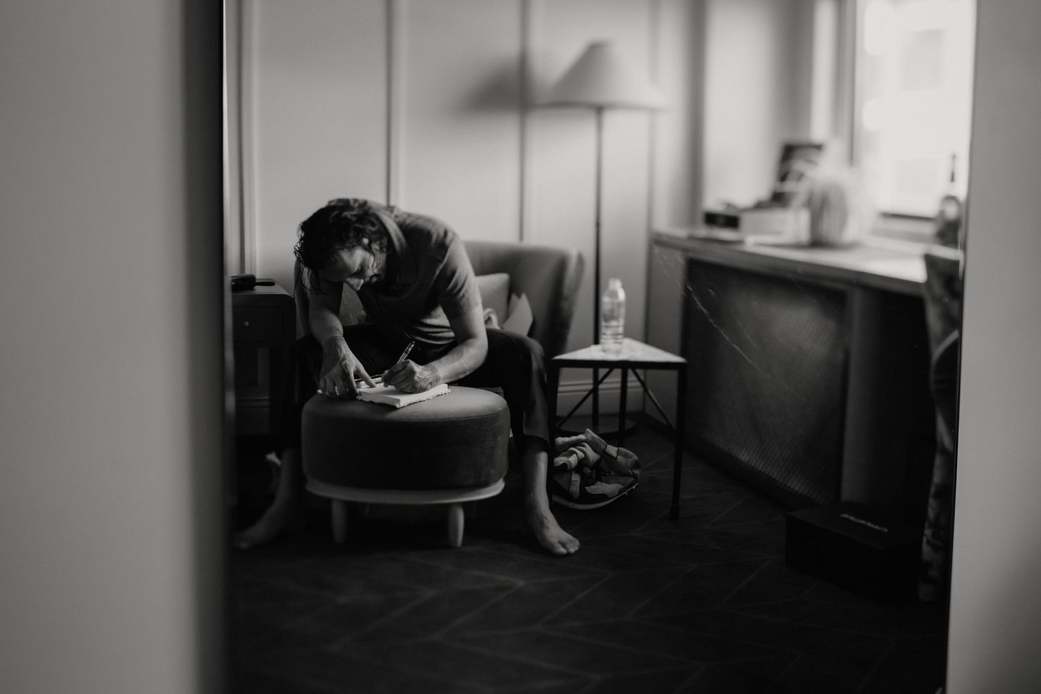 Black and white photo of a person sitting alone on an ottoman in a dimly lit room, lost in thought.