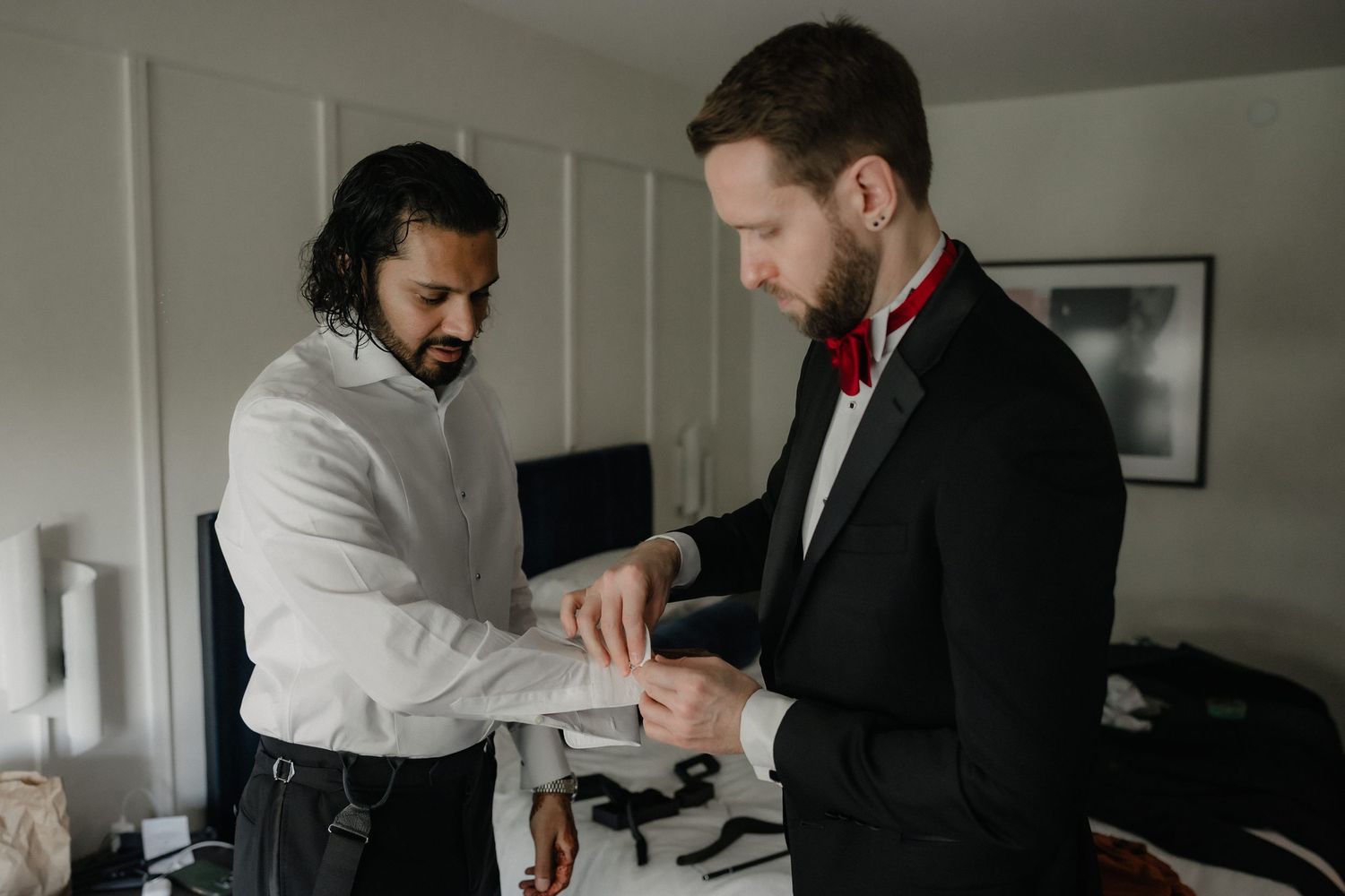 Groomsman helps groom adjust his cufflinks while preparing for the wedding ceremony indoors.