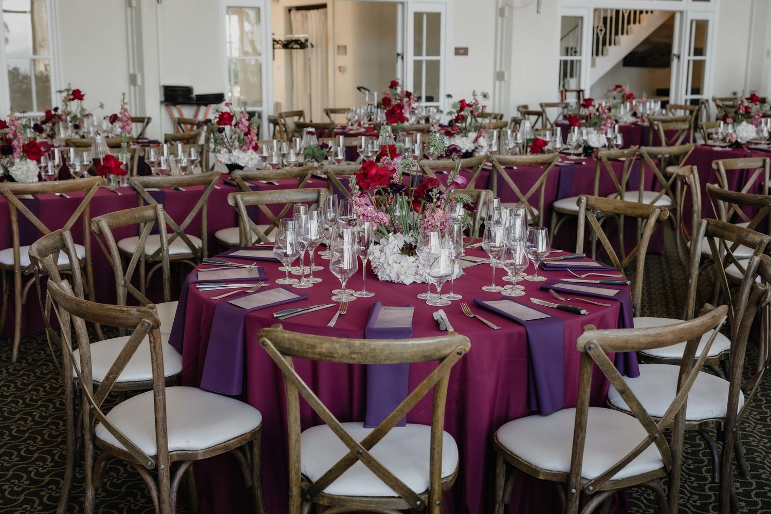 Elegant wedding reception with magenta tablecloths, rustic wooden cross-back chairs, and red floral centerpieces in a bright ballroom.