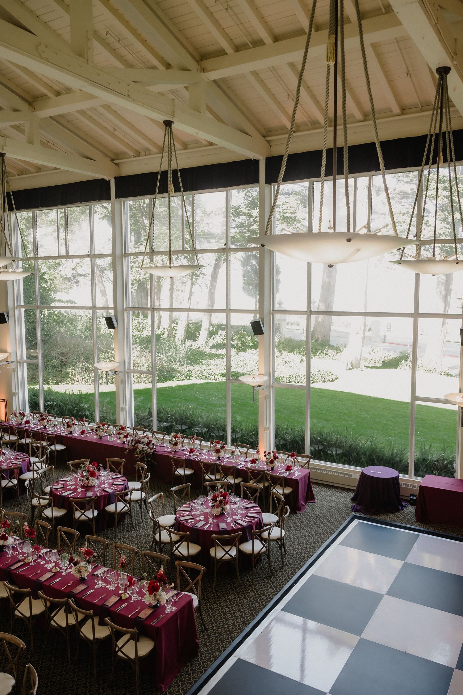 Elegant wedding reception hall with magenta tablecloths, round tables, and large windows overlooking a garden.