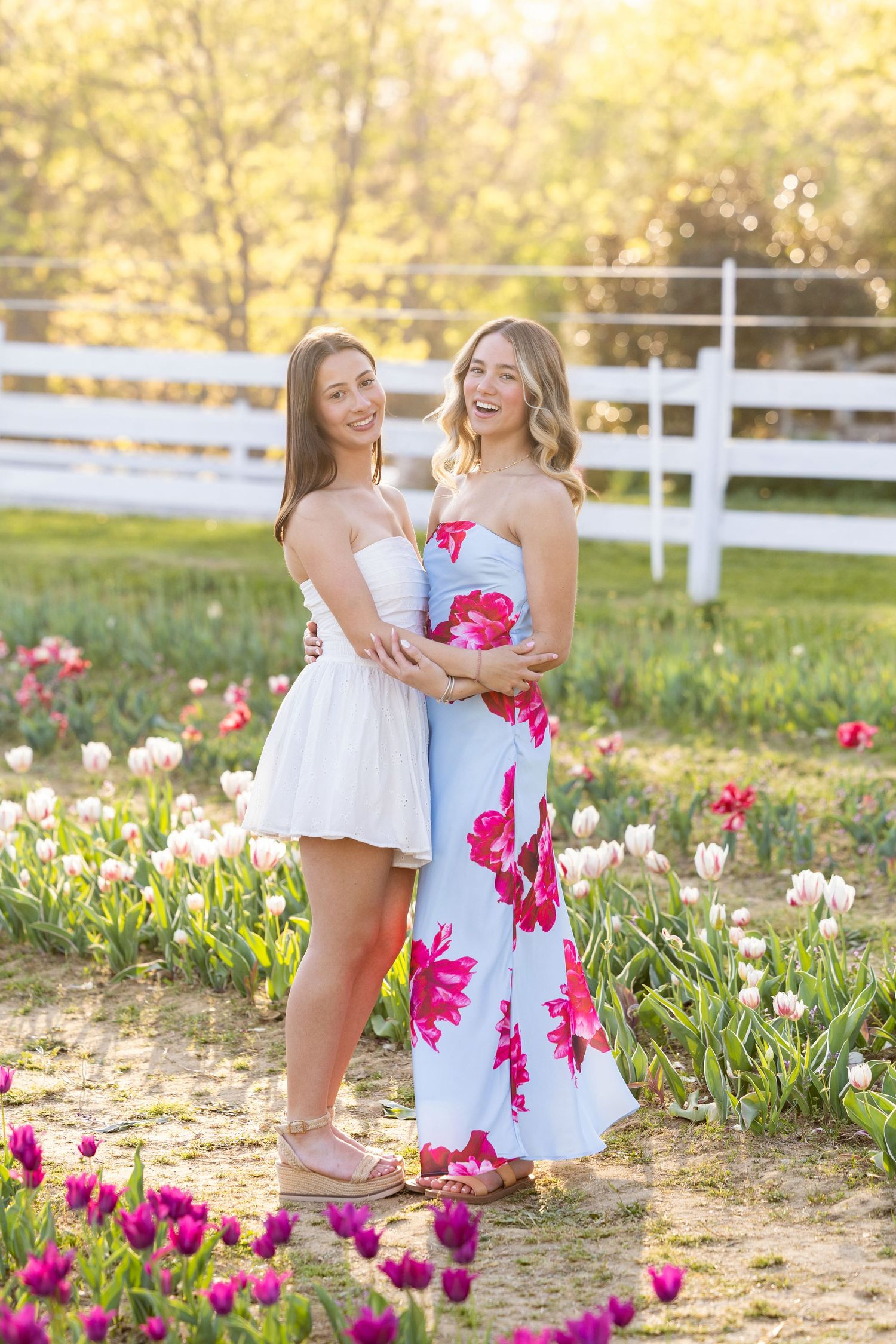 Two young women in summer dresses pose among colorful tulips in a sunny garden with a white fence.