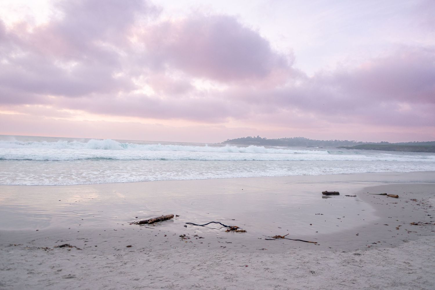 Serene beach at sunset with soft pink and purple clouds reflecting on wet sand and gentle ocean waves.