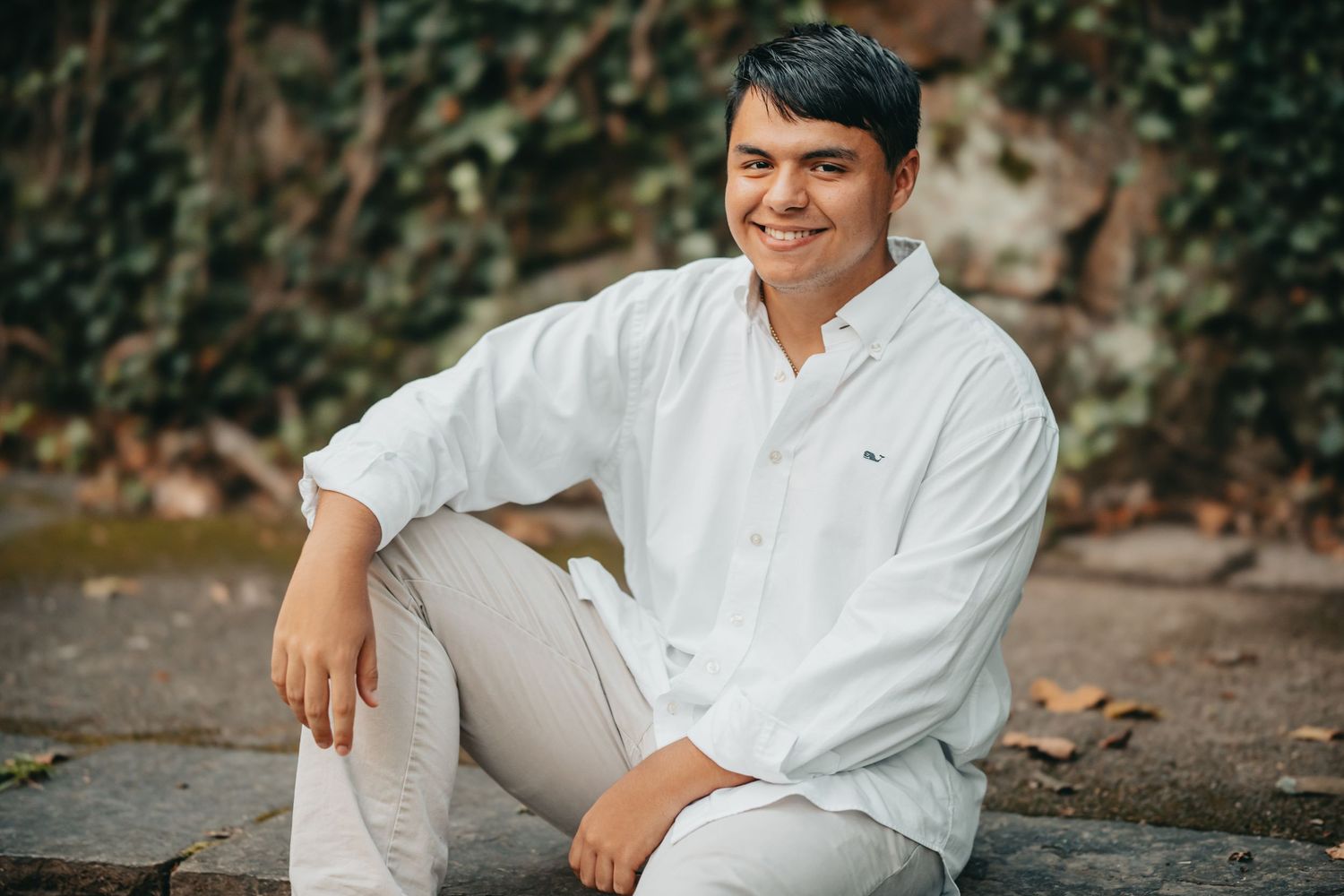 Young man in white Lacoste shirt and khakis sitting outdoors on stone steps, smiling confidently.