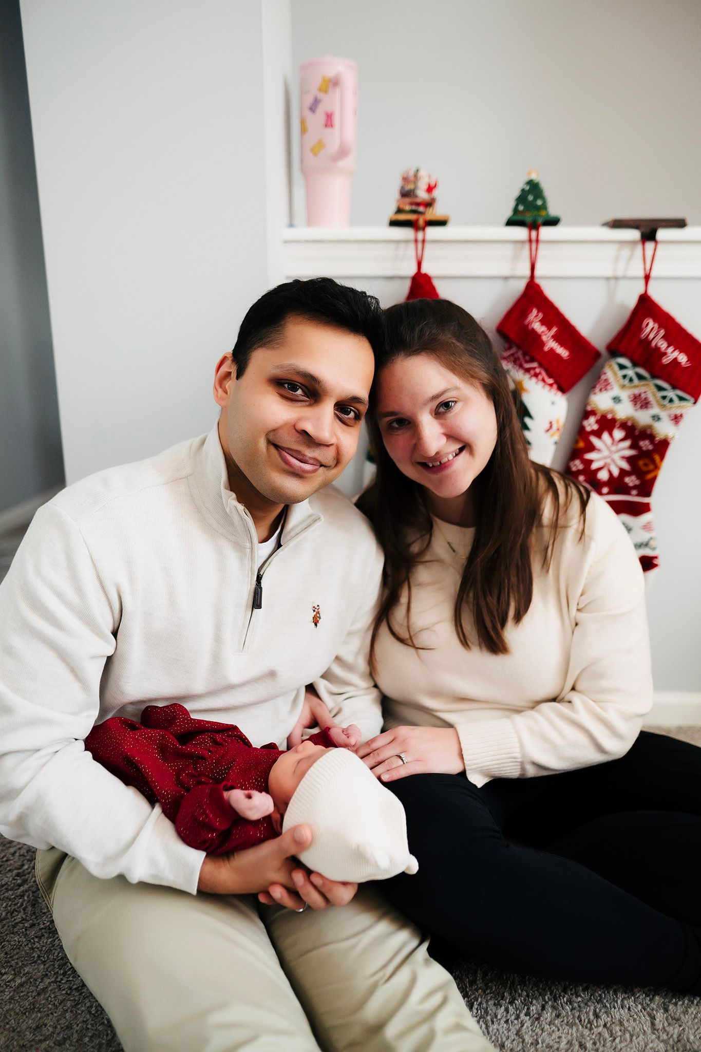 A couple sits together holding their newborn baby in front of a Christmas fireplace mantel with holiday stockings.
