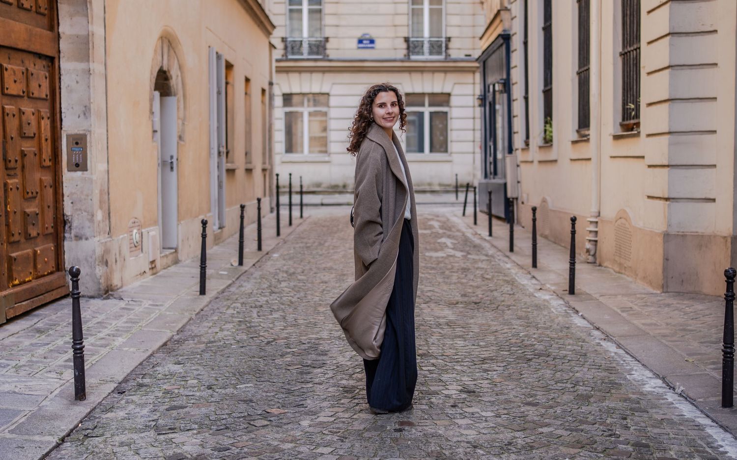 Woman in gray coat and navy pants posing on a cobblestone street in Paris, France.