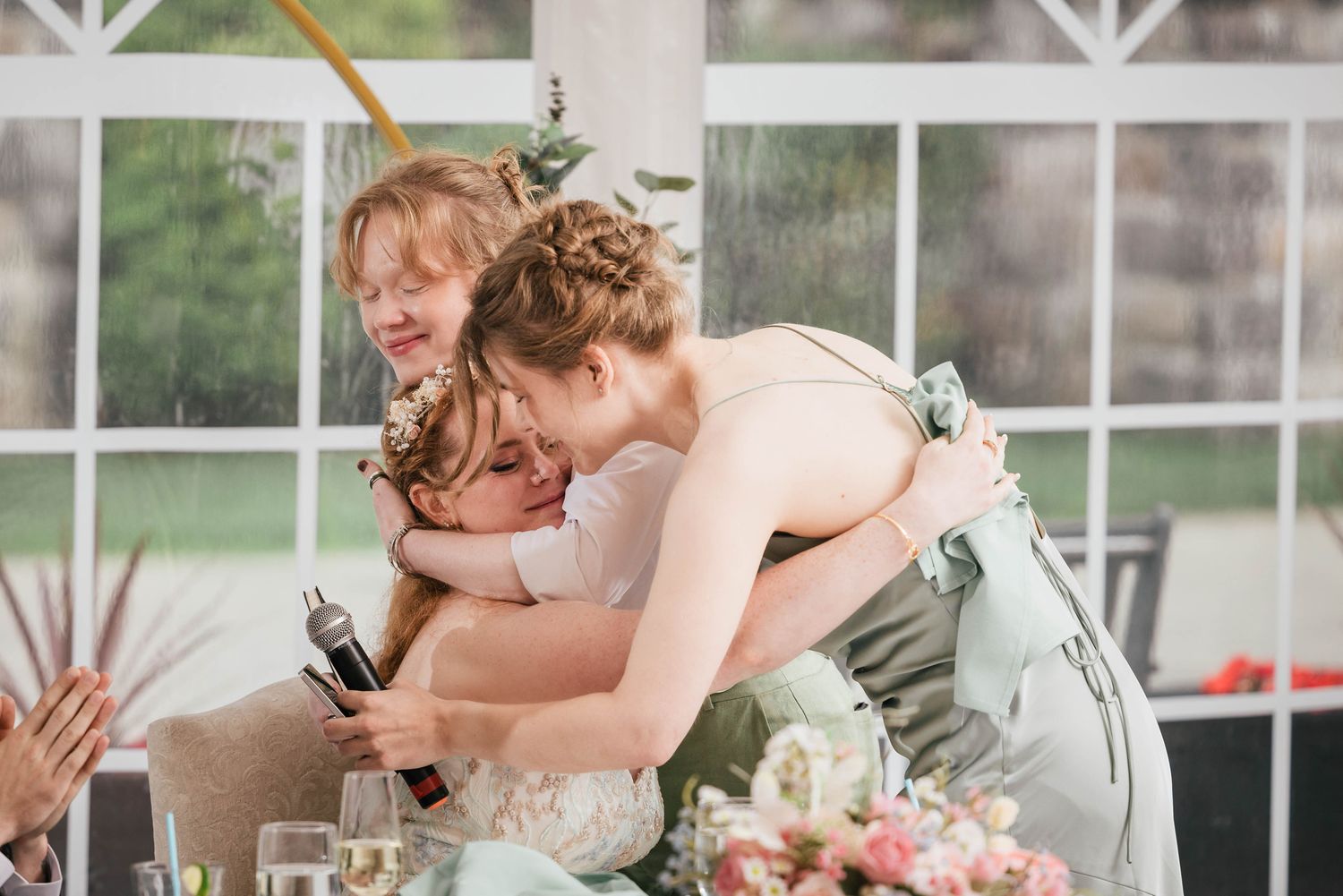 Bride embracing two bridesmaids in a tearful group hug after a speech at the reception.