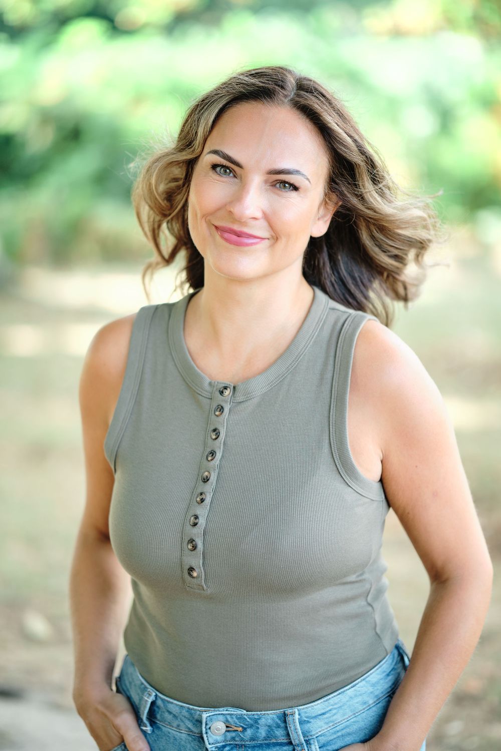 Smiling woman in olive green sleeveless henley top and jeans posing outdoors with blurred green foliage background.
