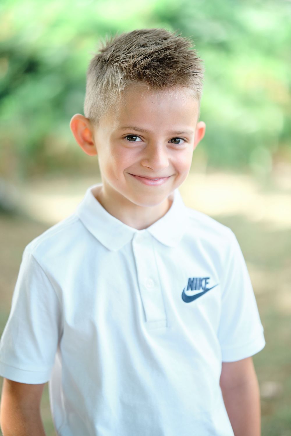 Smiling young boy wearing a white Nike polo shirt outdoors with green trees in the background.