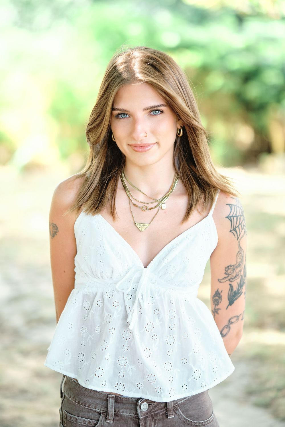 Young woman with blue eyes and tattoos wearing a white eyelet top and layered gold necklaces, smiling outdoors.