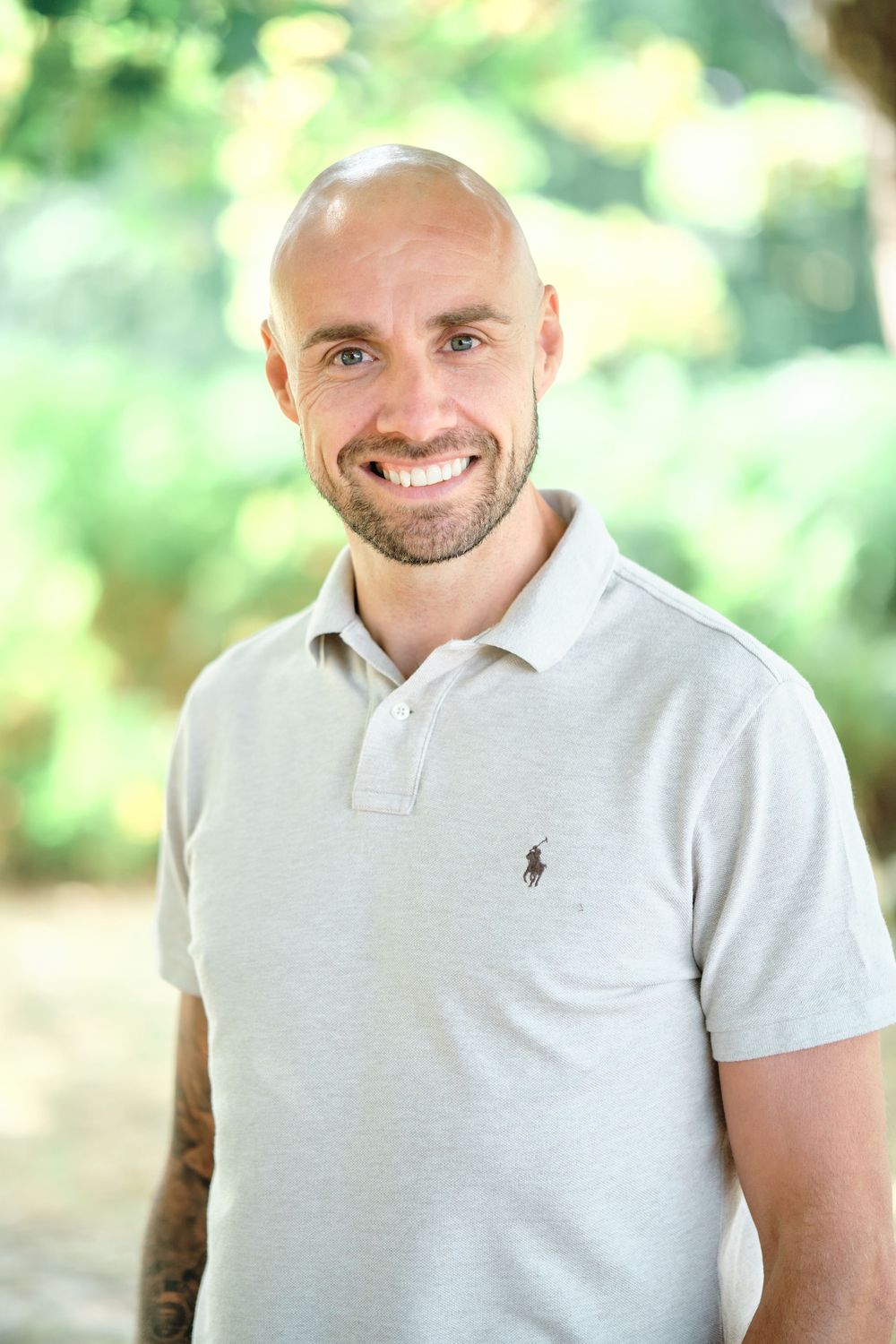 Smiling bald man with beard wearing a gray Ralph Lauren polo shirt outdoors with green trees in background.