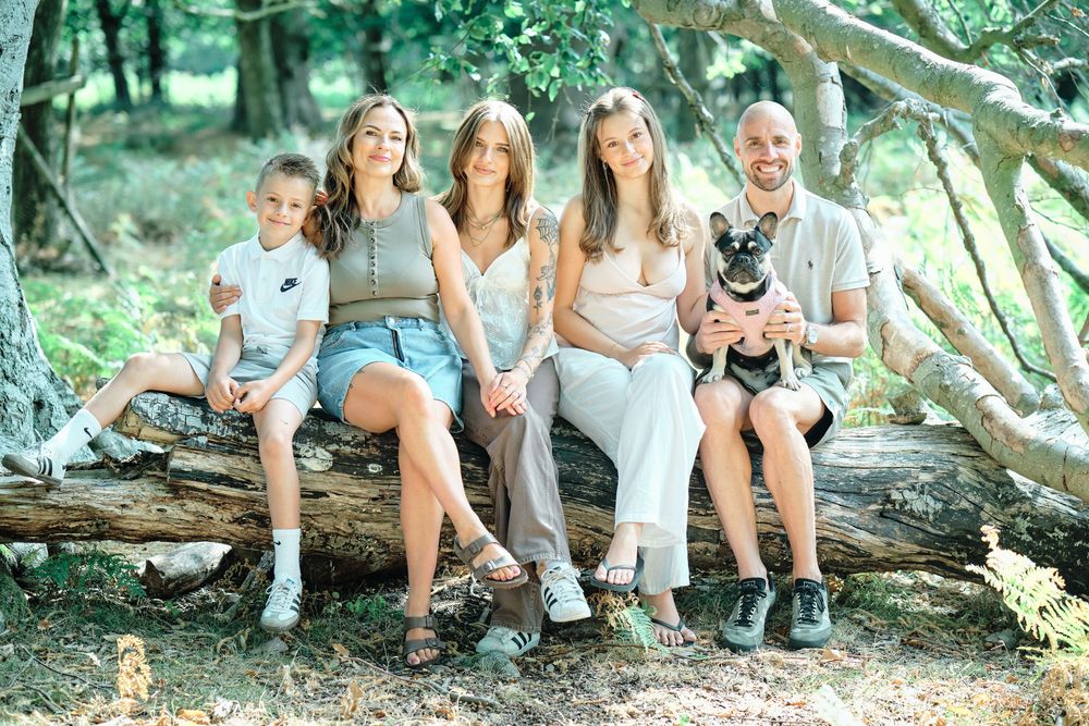 Family of four with a dog sitting on a fallen log in a lush green forest for an outdoor portrait.