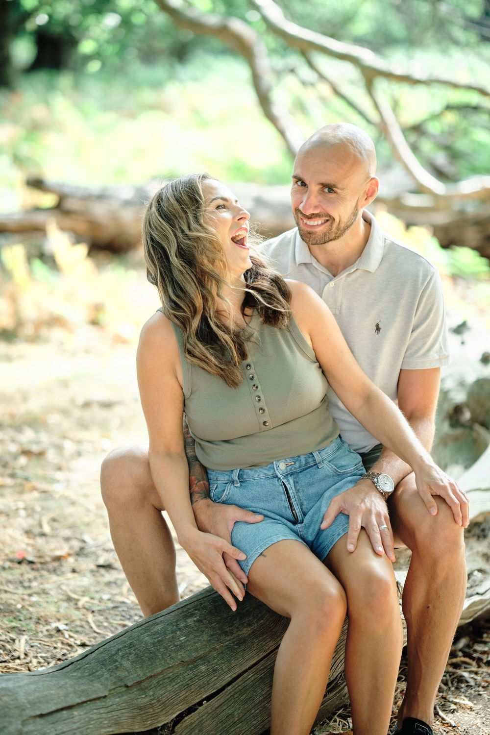 A laughing couple sits on a log in a forest, woman in sage crop top and denim shorts, man in white polo with a watch.