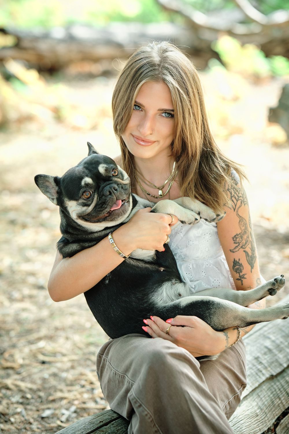 Young woman with tattoos holds a black French Bulldog outdoors in a wooded setting.