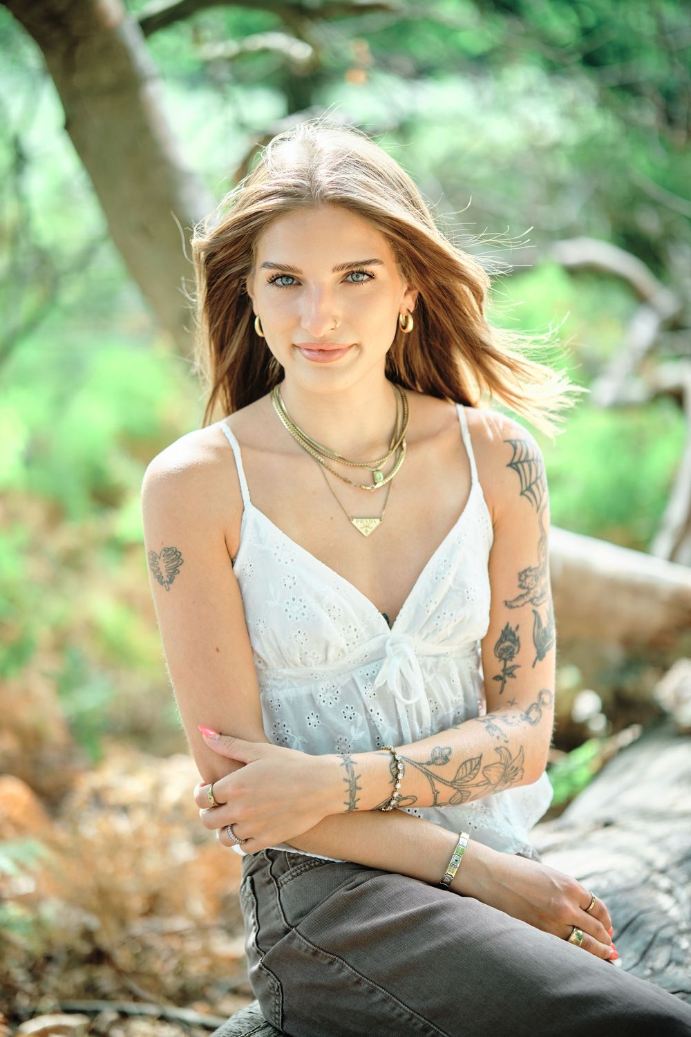 Young tattooed woman in white camisole and gold jewelry poses outdoors in a lush green forest setting.