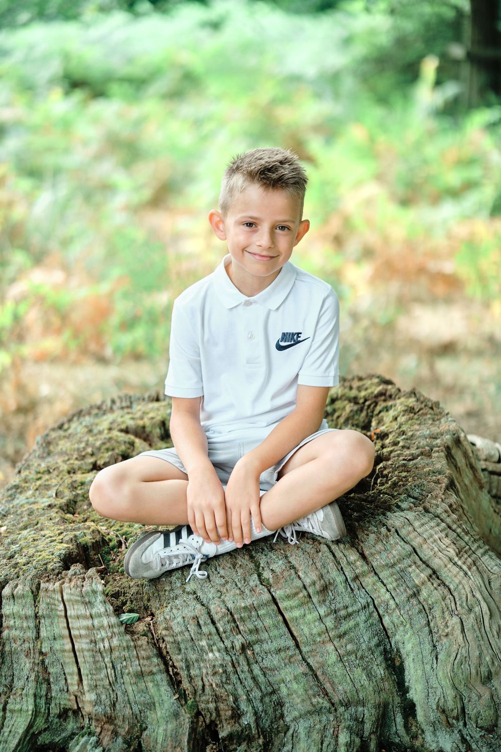 Smiling boy in white Nike polo shirt sitting cross-legged on a large mossy tree stump in a forest.